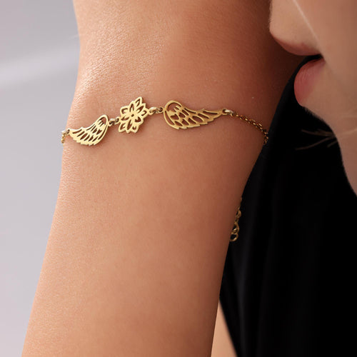 Close-up of a gold-toned bracelet featuring a delicate birth flower charm flanked by two angel wings, worn on a person's wrist.