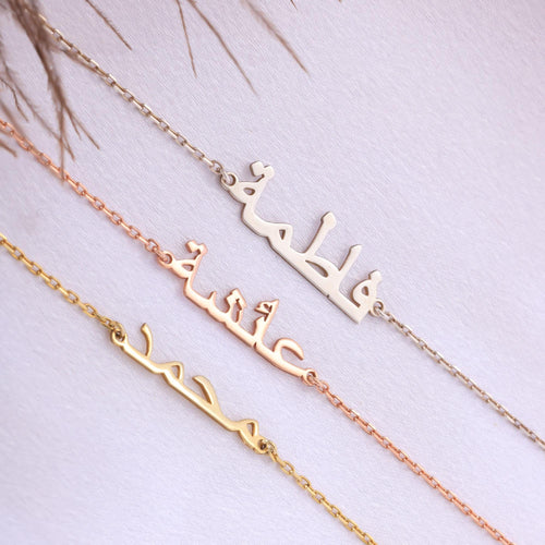 May include: Three name necklaces in gold, rose gold, and silver. Each necklace features a chain with a name written in Arabic script. The names are 'Fatima', 'Aisha', and 'Mohamed'. The necklaces are displayed on a white surface.