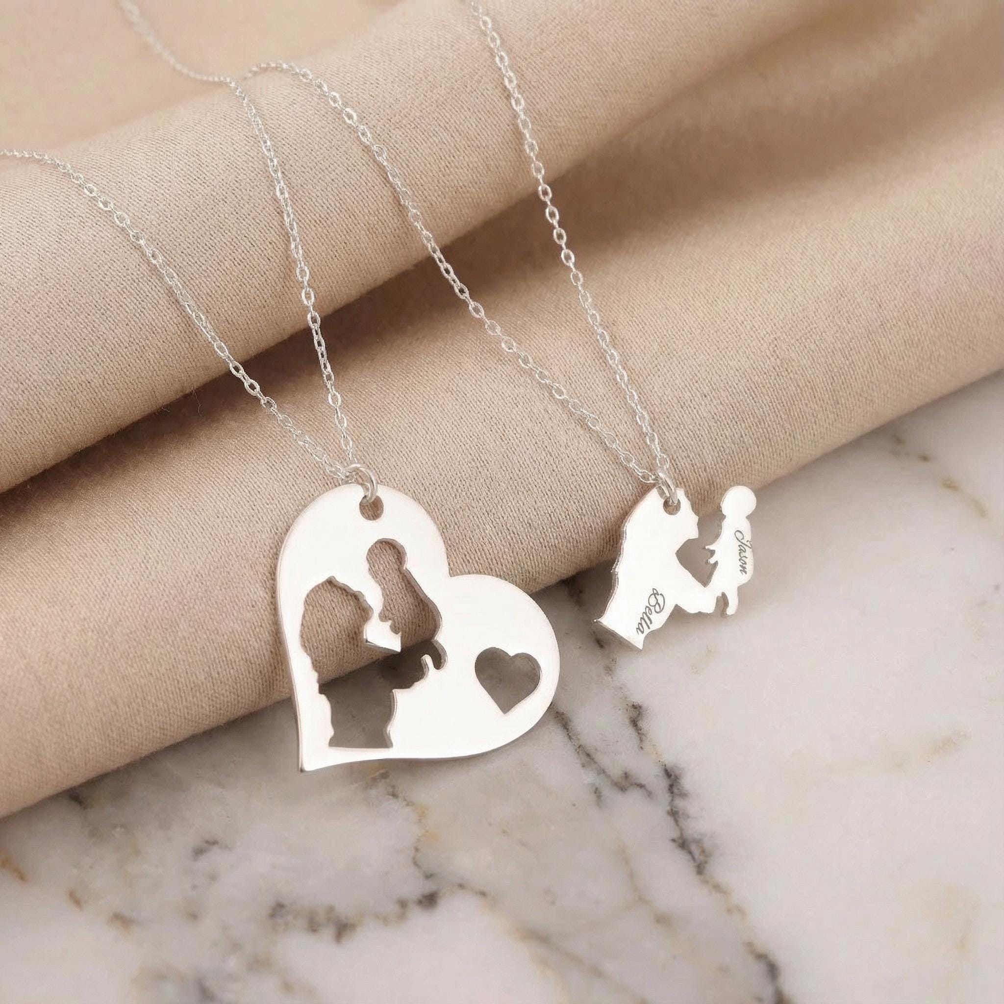 May include: Two silver necklaces with pendant charms. One pendant is a heart shape with a cutout of a couple. The other pendant is a silhouette of a person holding a child. The names "Bella" and "Jason" are engraved on the pendants.