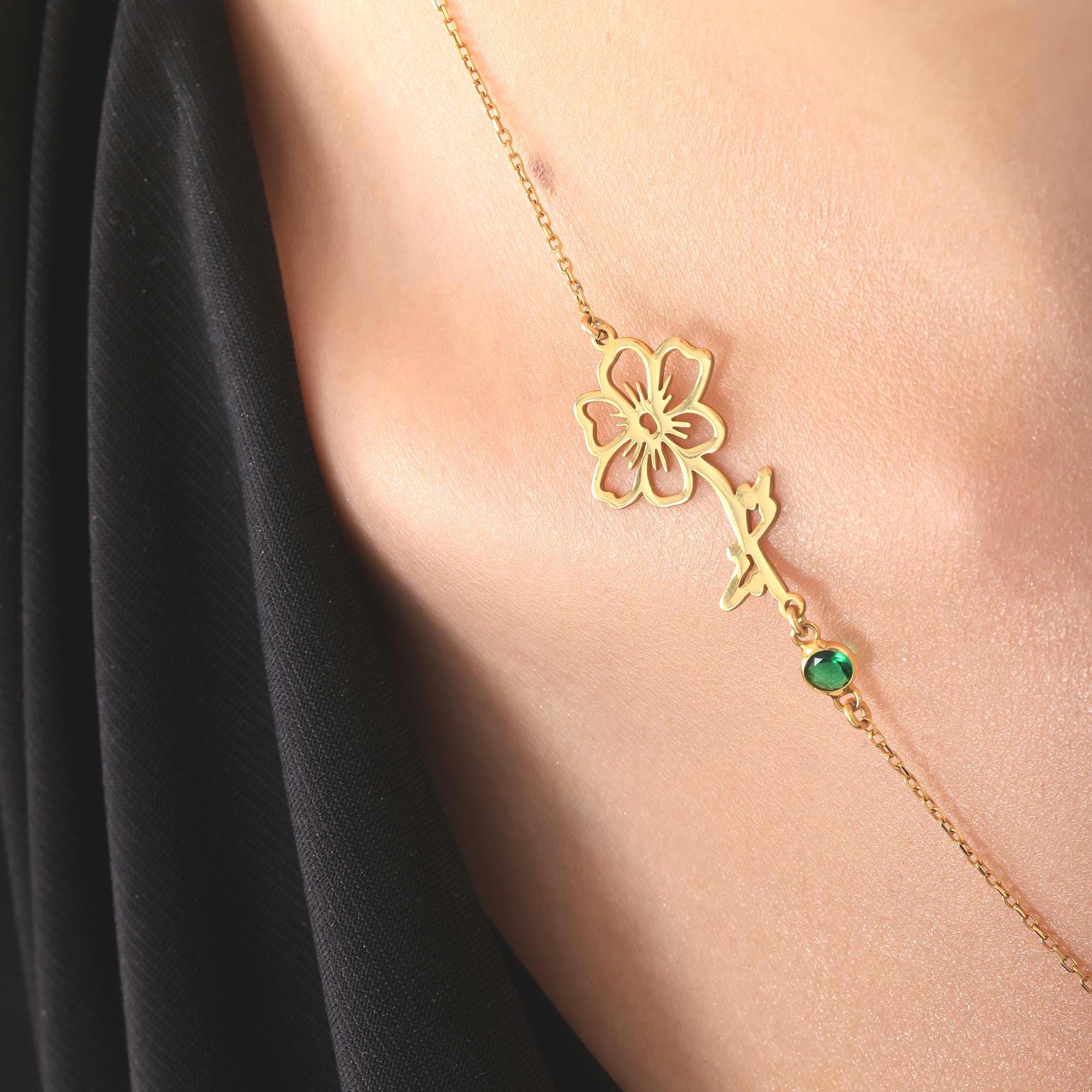 A gold chain necklace with a delicate flower pendant and a green gemstone accent. The flower pendant is a cutout design with a stem and leaves.