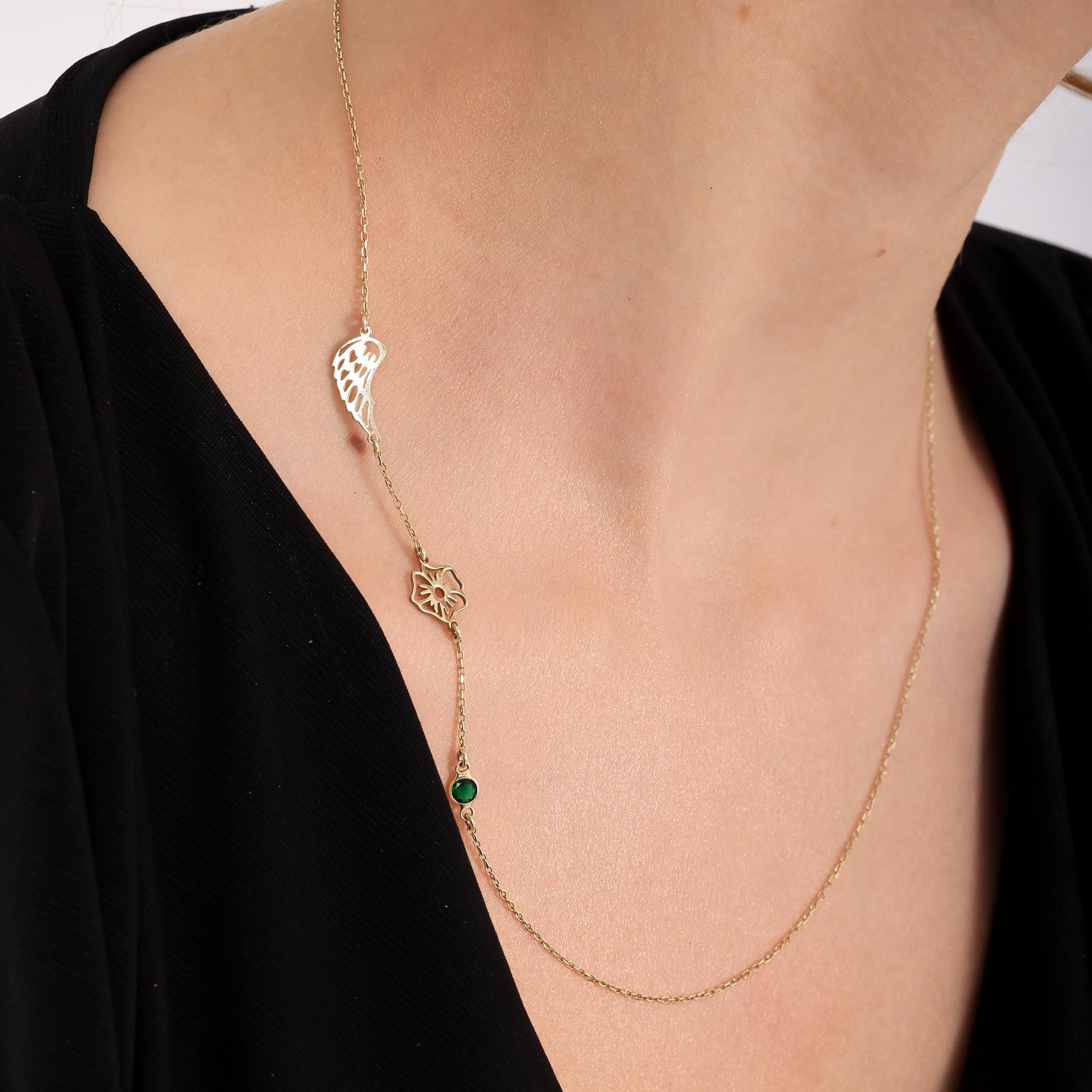 May include: A delicate gold necklace featuring three charms. The charms include a wing, a flower, and a small green gemstone. The necklace is displayed against a dark background, highlighting the gold color and intricate details of the charms. The necklace is a piece of jewelry.
