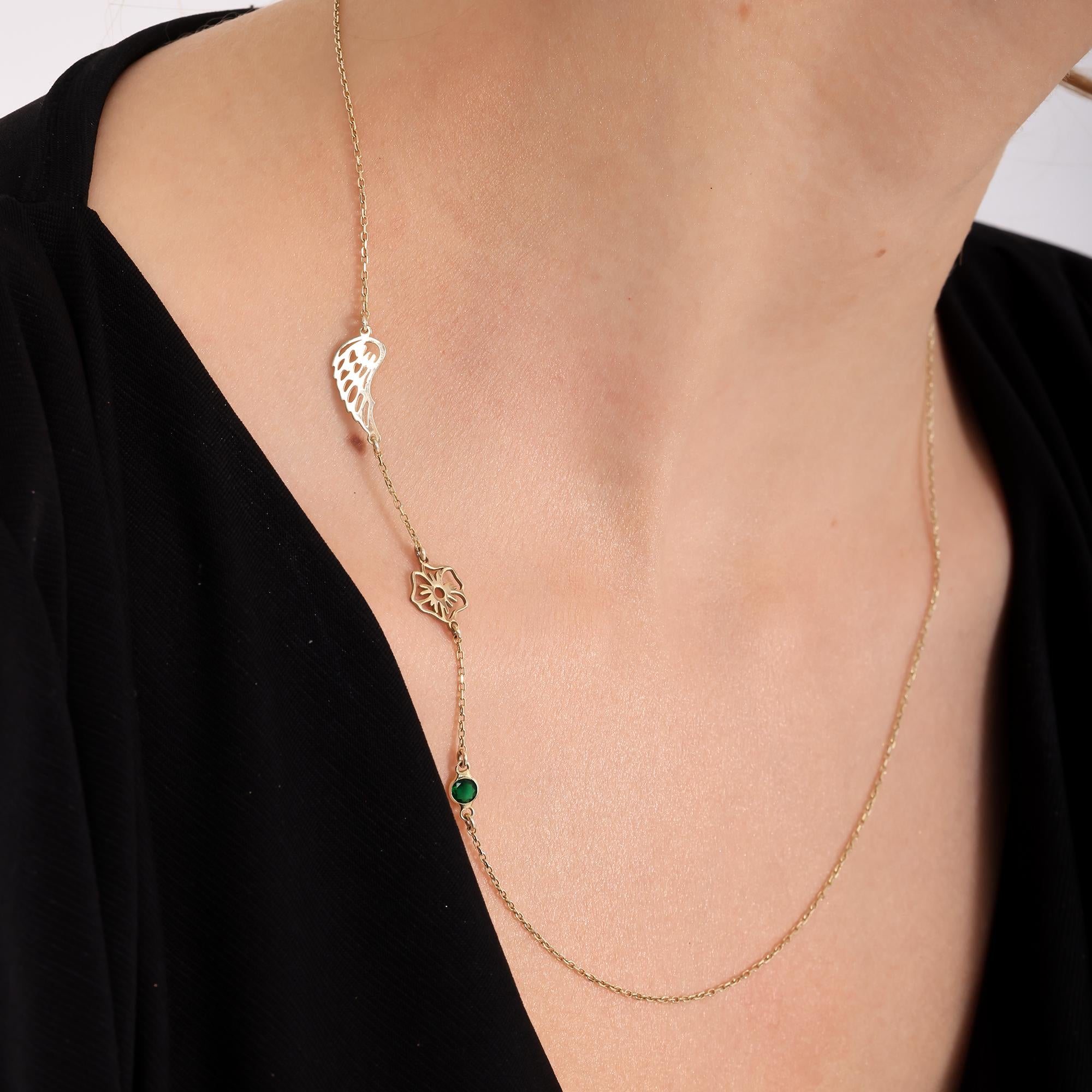 May include: A delicate gold necklace featuring three charms. The charms include a wing, a flower, and a small green gemstone. The necklace is displayed against a dark background, highlighting the gold color and intricate details of the charms. The necklace is a piece of jewelry.
