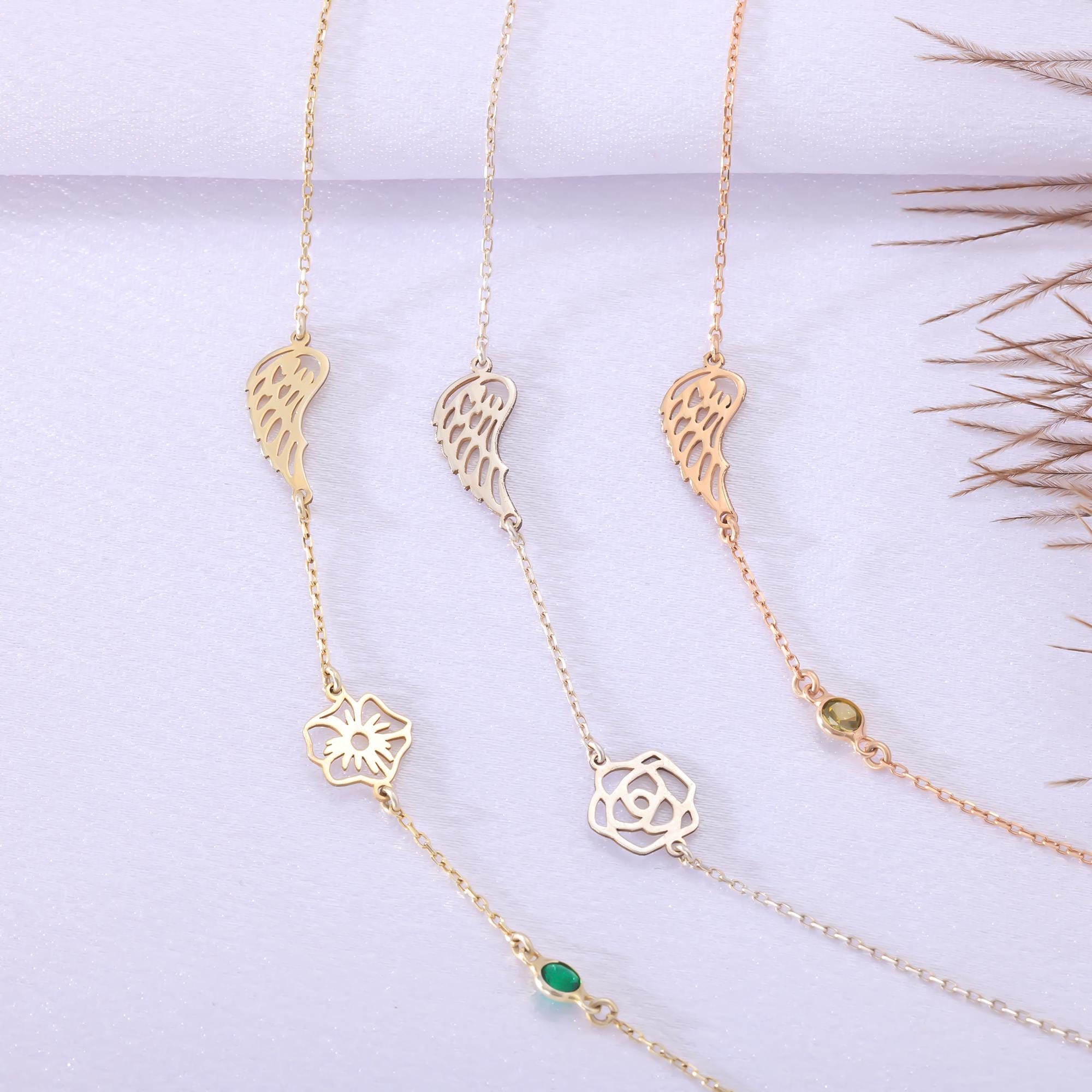 A flat lay display of three delicate necklaces. Two gold-toned necklaces each feature an angel wing charm and a unique birth flower design, both accented with a green birthstone. The third necklace is silver-toned, showcasing an angel wing, a different birth flower, and a purple birthstone.