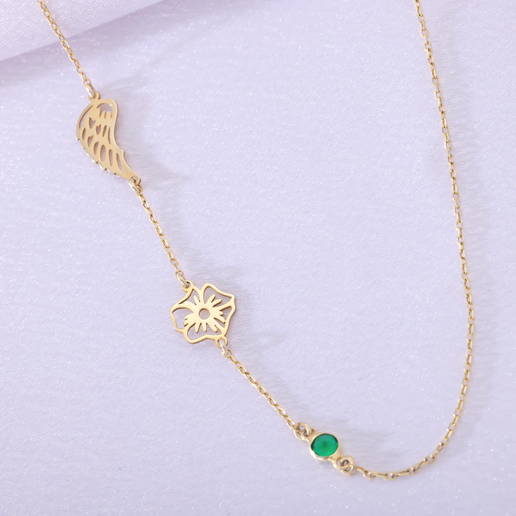 Close-up of a delicate gold-toned necklace featuring an angel wing charm, a birth flower charm, and a small green gemstone, lying on a light surface.