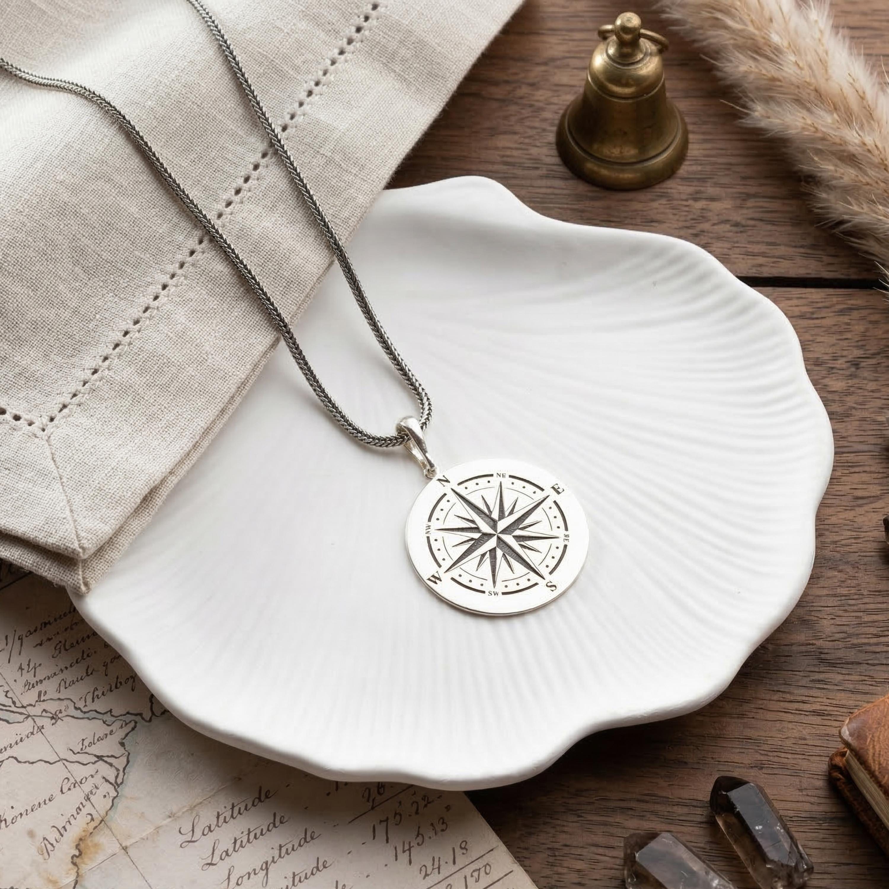 May include: A silver compass pendant necklace with a detailed compass rose design. The pendant hangs on a silver chain and rests on a white, shell-shaped dish. The necklace is a nautical-themed accessory.