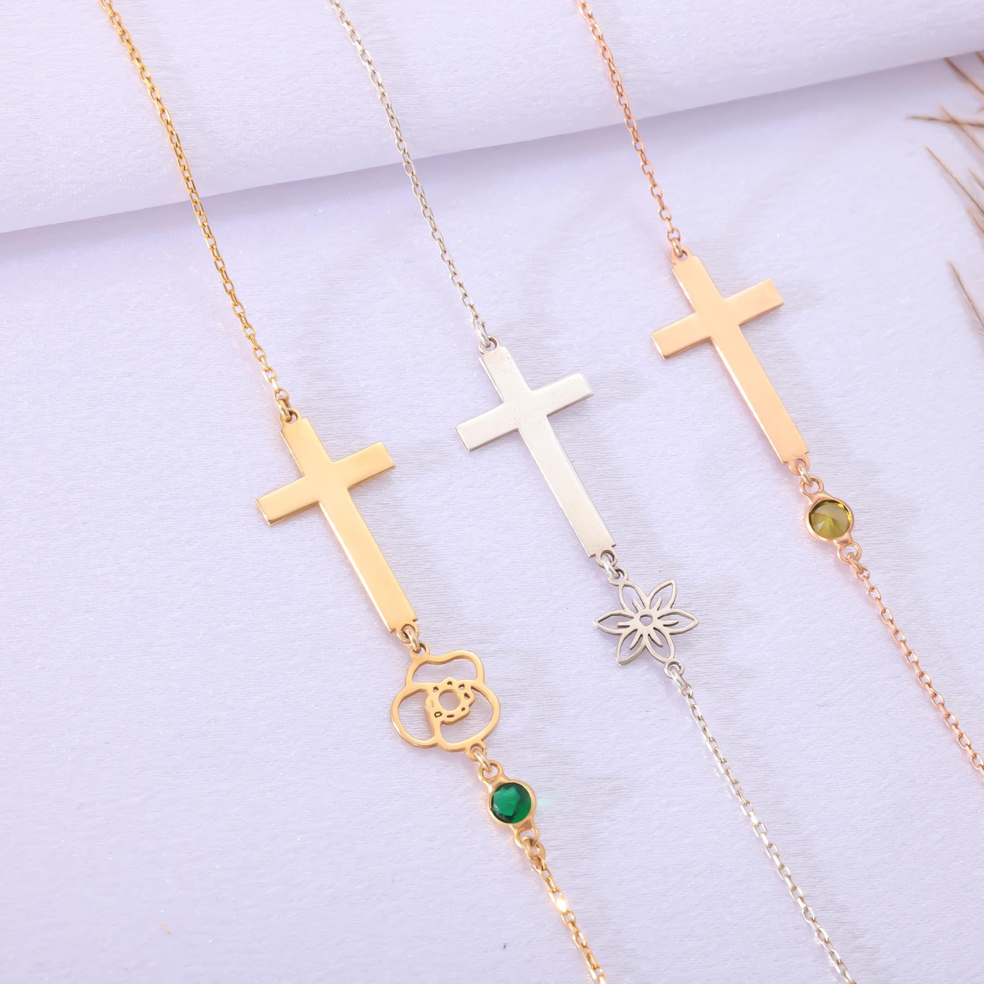 May include: Three gold, silver, and rose gold necklaces with cross pendants. Each necklace has a different charm hanging from the cross. The gold necklace has a green gemstone and a flower charm. The silver necklace has a flower charm. The rose gold necklace has a green gemstone.