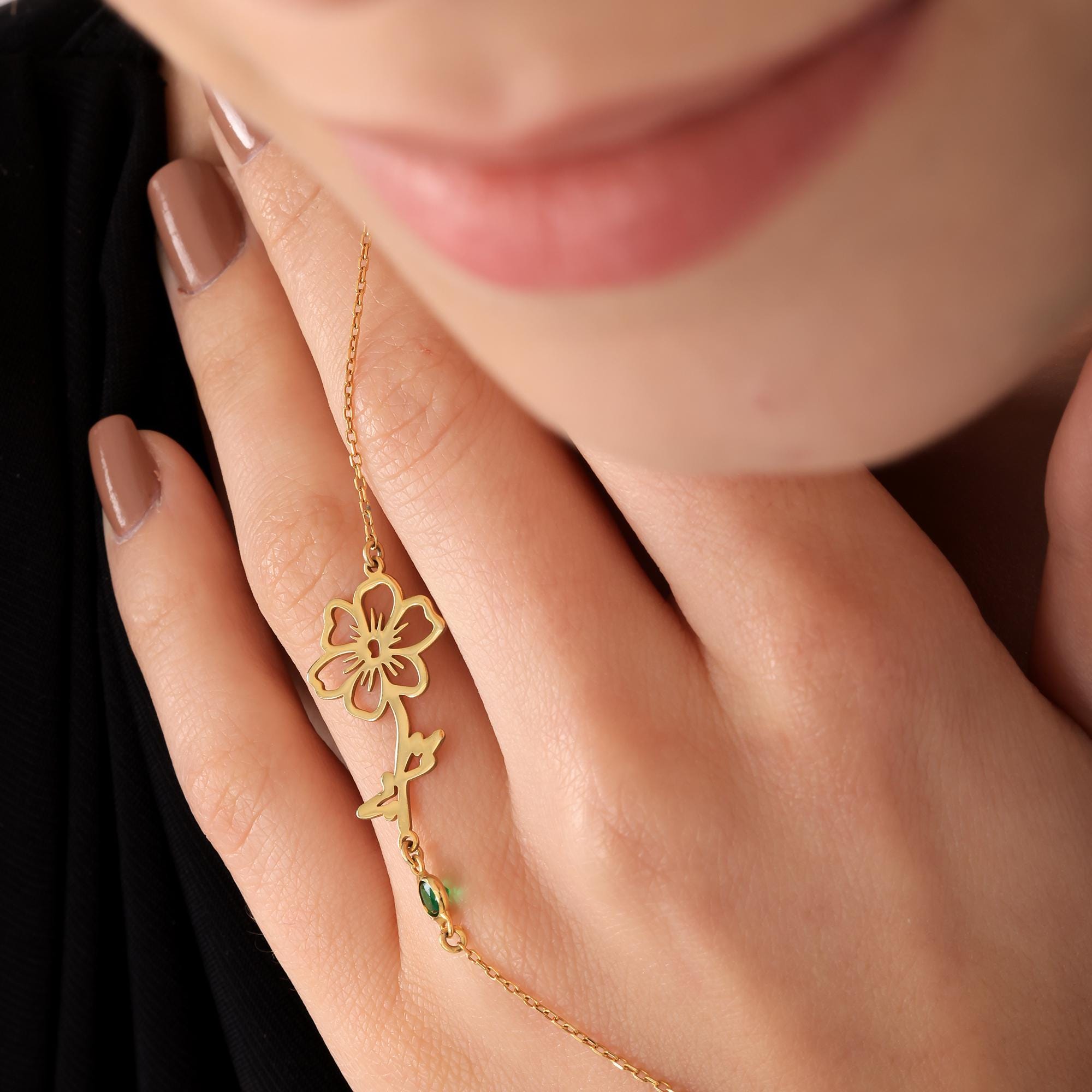 A gold flower pendant necklace with a green gemstone. The pendant is in the shape of a flower with a stem and leaves.