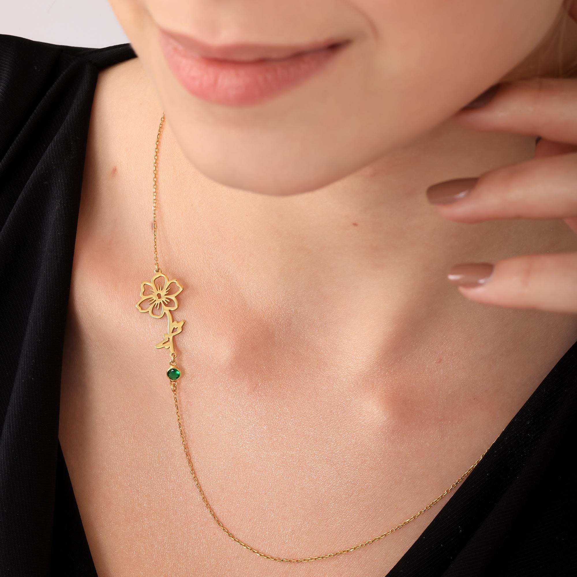 A gold chain necklace with a delicate flower pendant and a green gemstone. The pendant is a cutout design of a flower with a stem and leaves.